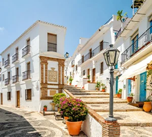 Calle empedrada con casas blancas, escalones, macetas con flores y farol. Cielo despejado.
