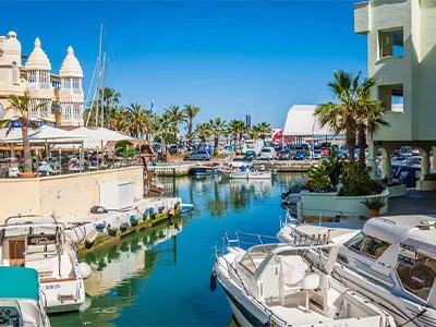 Puerto con barcos, palmeras y edificios junto al agua, cielo despejado.