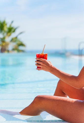 Persona sosteniendo cóctel junto a una piscina bajo el sol.