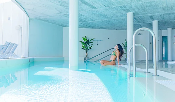Mujer sentada en el borde de una piscina interior, con plantas al fondo.