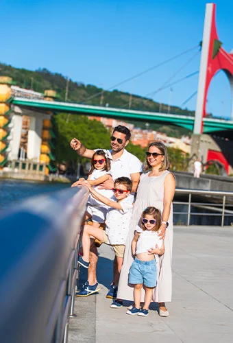 Familia con gafas de sol sonríe junto a una barandilla, con un puente de fondo.