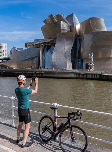 Persona en bici frente al Museo Guggenheim Bilbao, tomando fotos junto al río en un día soleado.