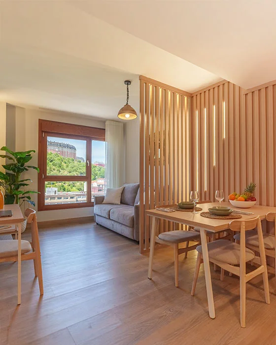 Sala con comedor moderno, sofá gris, ventana amplia y decoración de madera clara.