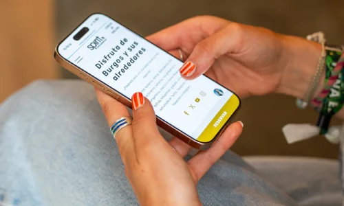 Persona sosteniendo un smartphone con texto sobre Burgos en la pantalla. Lleva anillos y pulseras.