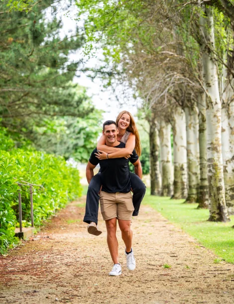Pareja sonriendo; hombre cargando a mujer en sendero rodeado de árboles.