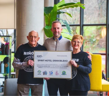 Tres personas sostienen un gran cheque del Spirit Hotel Gran Bilbao en un entorno interior.