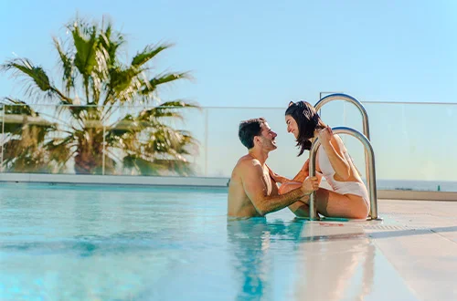 Pareja sonriendo en una piscina bajo un cielo despejado, junto a una palmera.