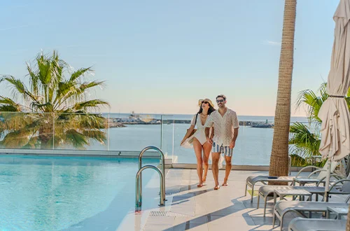 Pareja junto a una piscina con vista al mar, rodeada de palmeras.