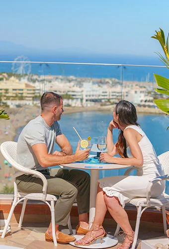 Pareja sentada en terraza con vista al mar y bebidas sobre la mesa.