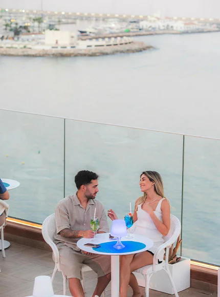 Pareja disfrutando bebidas en una terraza con vista al mar.