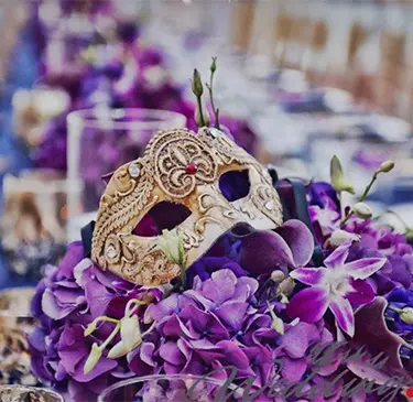 Máscara dorada sobre un arreglo floral púrpura con hojas verdes y velas alrededor.