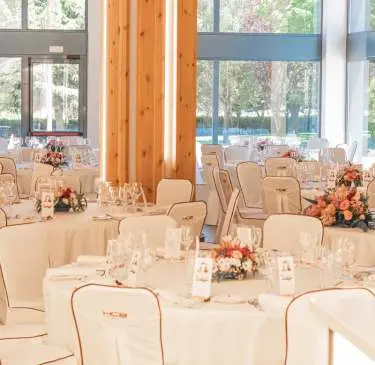 Salón de eventos con mesas redondas decoradas con flores y copas sobre manteles blancos.
