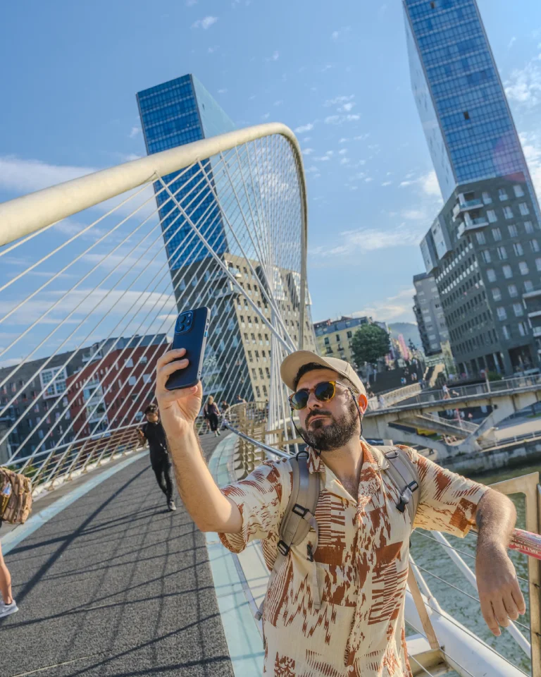 Persona tomando una selfie en un puente con edificios modernos de fondo.