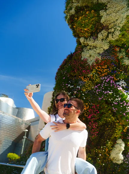 Pareja tomándose una selfie frente a una gran escultura floral bajo un cielo despejado.