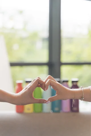 Dos manos formando un corazón delante de botellas de colores sobre una mesa.