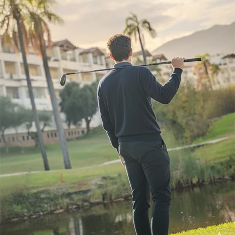 Persona con palo de golf en un campo, frente a una laguna y edificios al fondo.