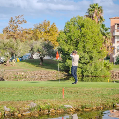Persona jugando golf en un campo verde con árboles y edificios al fondo, bajo un cielo azul.