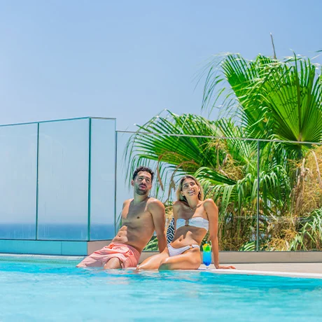 Pareja sentada al borde de una piscina con palmeras y cielo azul de fondo.
