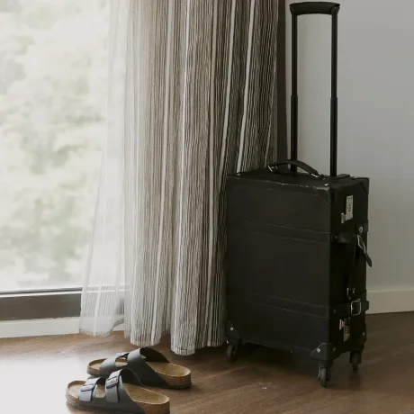 Una maleta negra junto a unas sandalias junto a una ventana con cortinas.