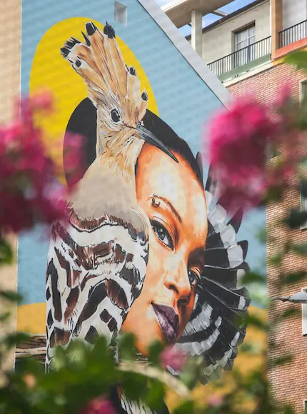 Mural de un rostro y un pájaro en la pared de un edificio, flores delante.