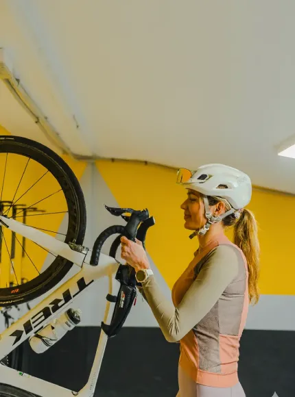 Mujer con casco ajustando una bicicleta en un interior de pared amarilla y gris.