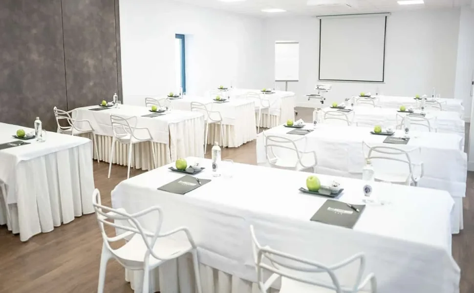 Aula de conferencias con mesas blancas, sillas modernas y pantalla de proyección.
