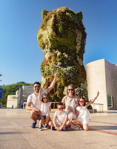 Familia sonriendo frente a la escultura de Puppy del Museo Guggenheim bajo un cielo despejado.