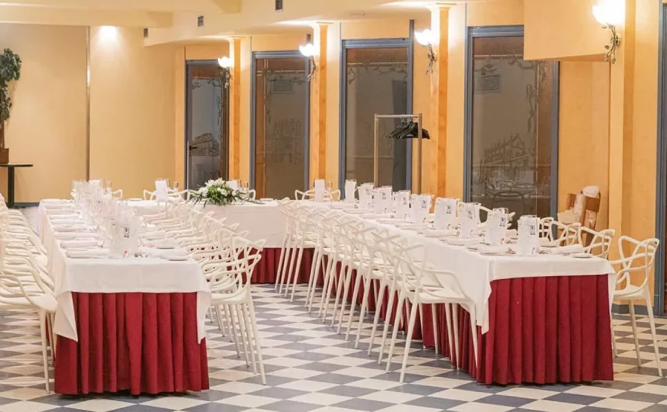 Salón de eventos con mesas largas vestidas con manteles blancos y faldones rojos.