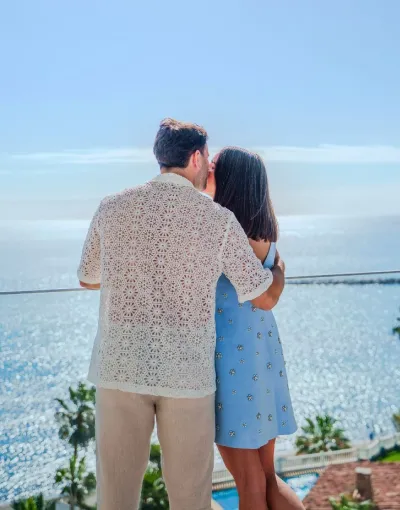 Pareja abrazada de espaldas contemplando el mar en un día soleado.