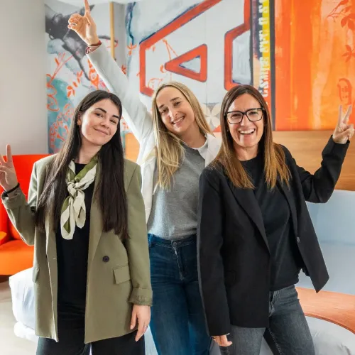 Tres mujeres sonrientes levantan los brazos en señal de victoria en una oficina colorida.