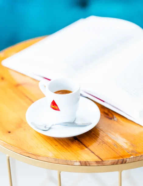 Taza de espresso con platillo y cuchara sobre mesa, libro abierto de fondo.