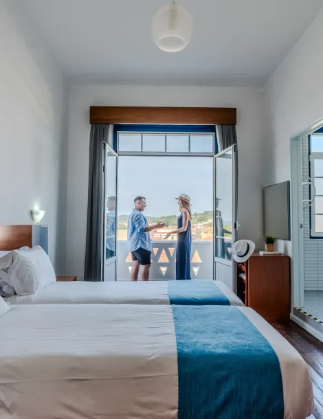 Habitación de hotel con dos camas; pareja de pie en el balcón disfrutando la vista al exterior.