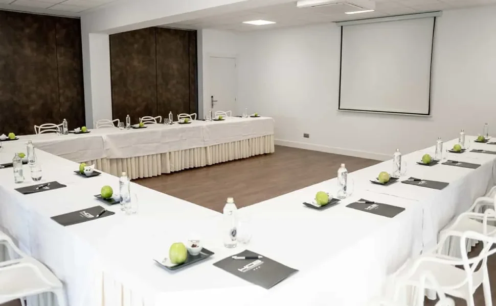 Sala de conferencias con mesas en forma de U, agua y manzanas. Pantalla al fondo.