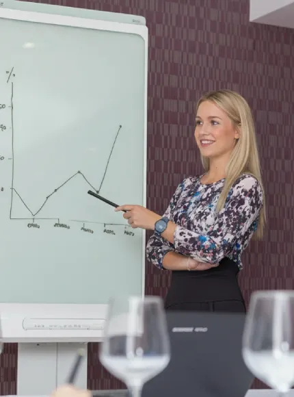 Mujer presentando un gráfico en una pizarra blanca, señalando con un puntero.