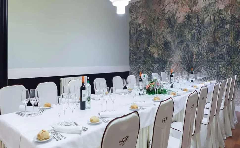Mesa elegante en restaurante, con vinos, copas y sillas blancas. Decoración vegetal.