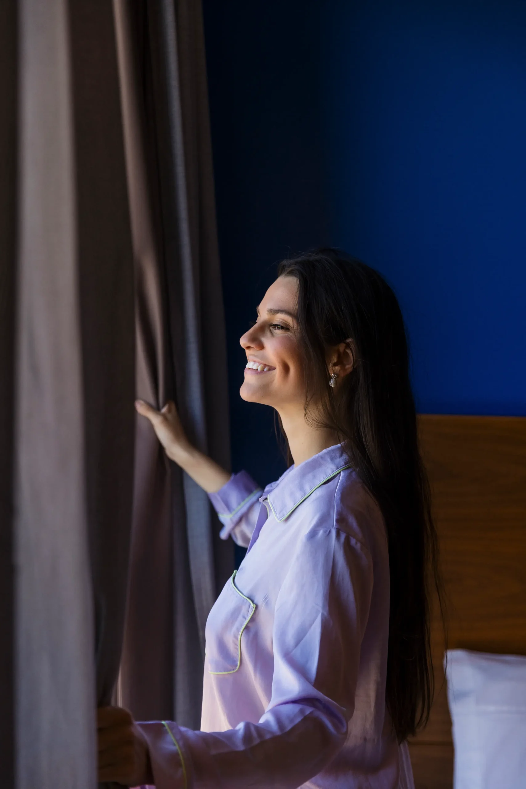Mujer sonriendo, abriendo cortinas al amanecer en una habitación azul.