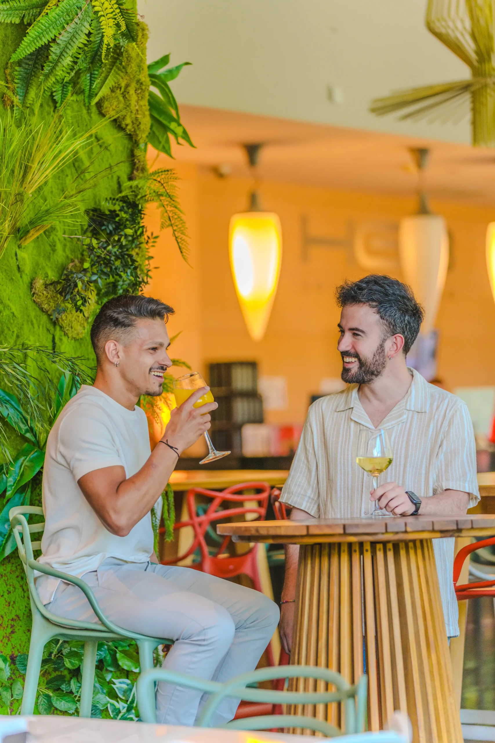 Dos hombres conversan y ríen con bebidas en un café con decoración vegetal.