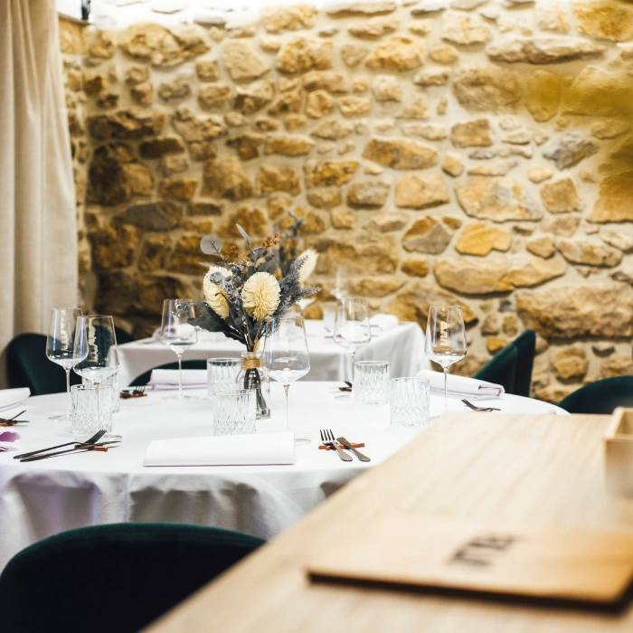 Mesa redonda elegante con mantel blanco, copas de vino, y un arreglo floral, frente a una pared de piedra rústica.