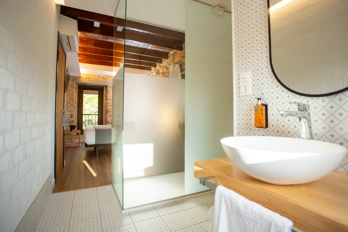 Baño moderno con lavabo de cerámica blanca, espejo ovalado y vista a un dormitorio con techo de madera y cama.