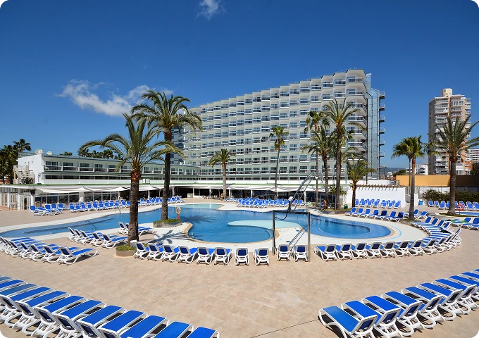 Piscina de hotel con sillones azules, palmeras y edificio moderno de fondo bajo un cielo despejado.