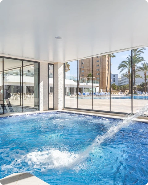 Piscina interior con cascada, al fondo se ve otra piscina exterior y palmeras.