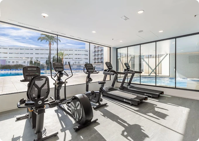 Gimnasio moderno con bicicletas estáticas y cintas de correr, junto a ventanas que dan a una piscina.