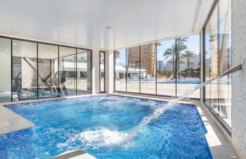 Piscina cubierta con chorros de agua y vista a palmeras a través de grandes ventanales.