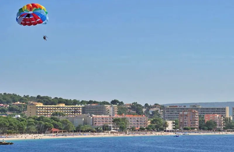 Paracaídas de colores sobrevuela una playa con edificios y mar calmo.