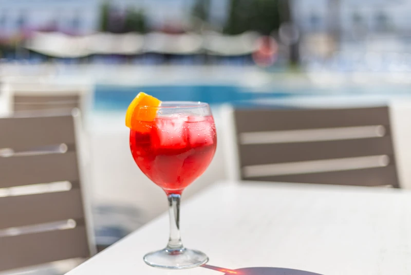Cóctel rojo con naranja y hielo en una mesa junto a una piscina.