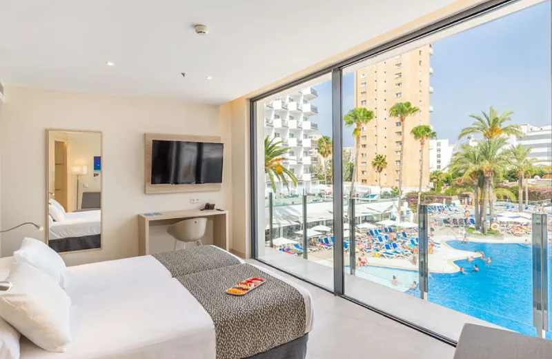 Habitación de hotel con vista a una piscina y edificios altos a través de un gran ventanal.