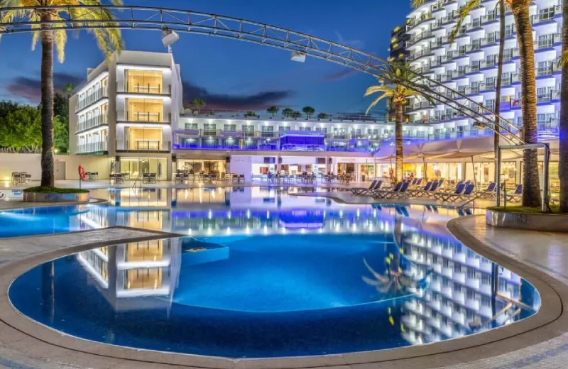 Hotel moderno con piscina iluminada al anochecer, rodeada de palmeras y tumbonas.