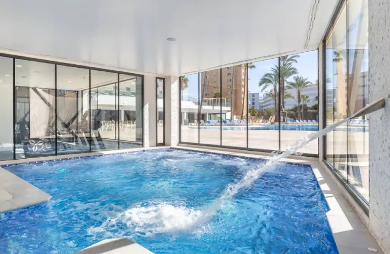 Piscina cubierta con ventanales y vista al exterior con palmeras y edificios.