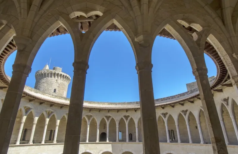Interior del castillo de Bellver con arcos góticos y torre de fondo bajo un cielo azul despejado.