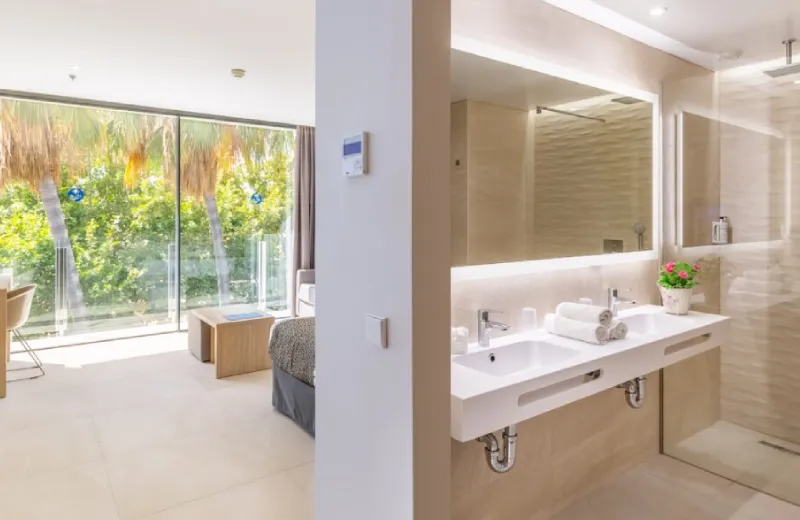 Habitación luminosa con baño integrado, cama, y ventanales con vista a palmeras.